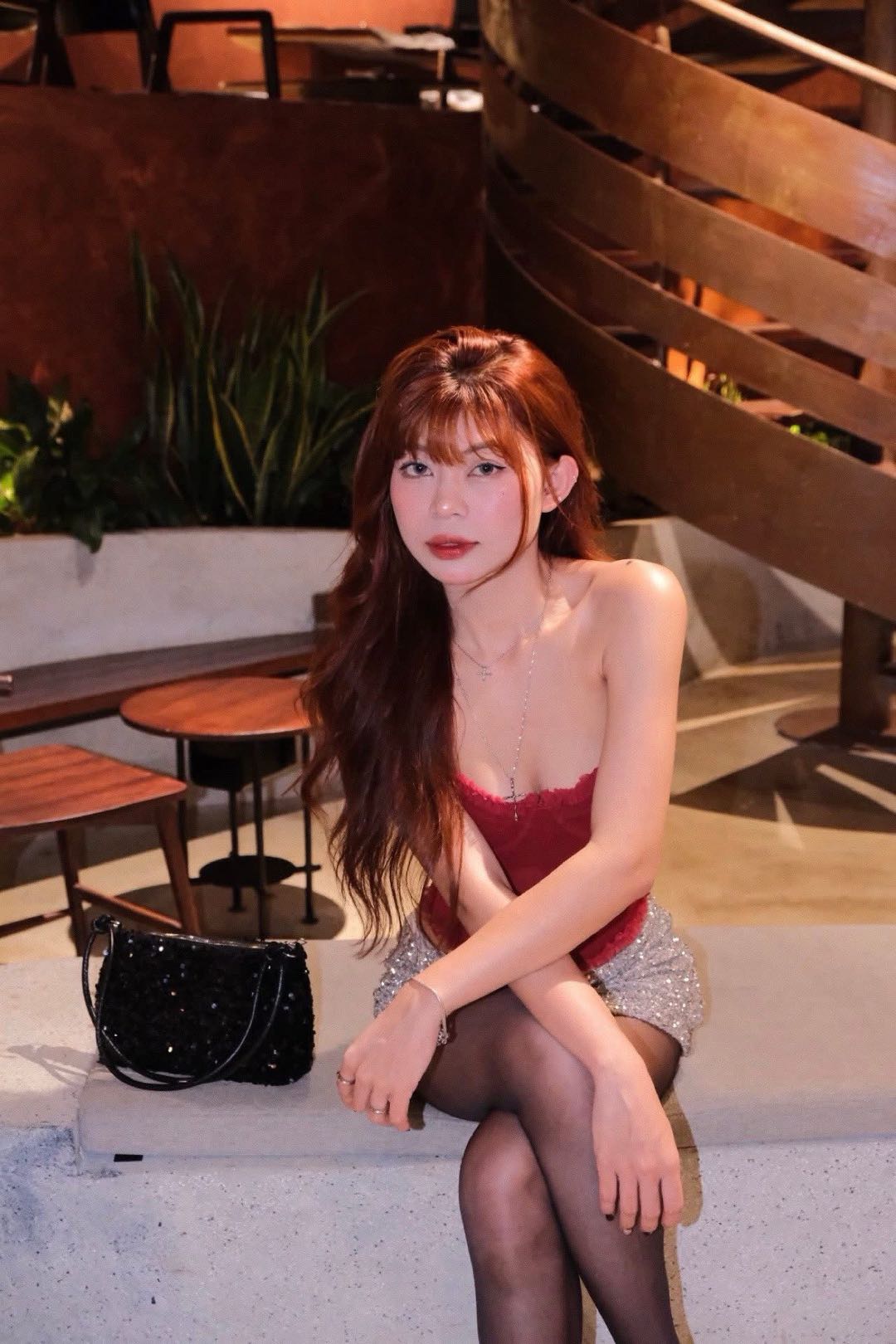A young woman from Vietnam with auburn hair and bangs, seated in a red corset top and sparkly skirt